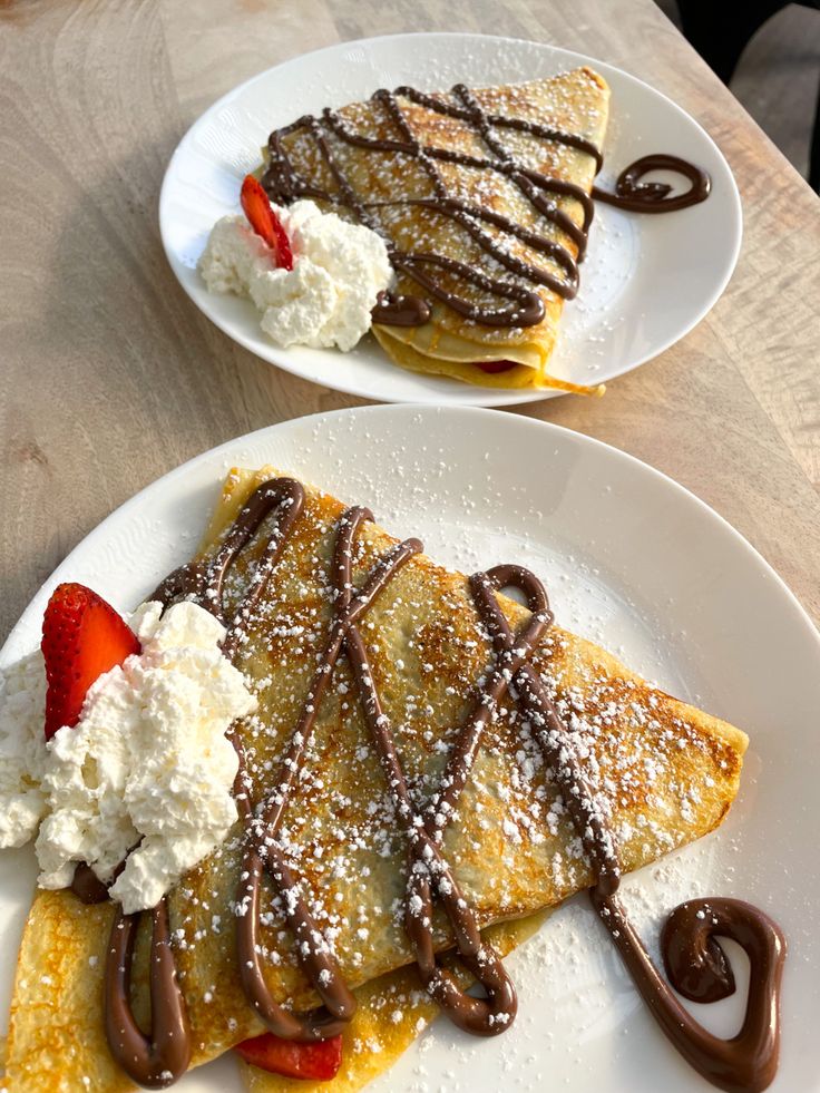 two white plates topped with crepes covered in chocolate and whipped cream next to each other