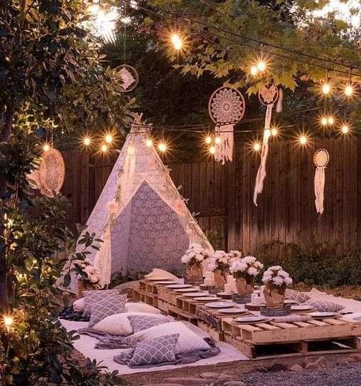 an outdoor dining area with lights strung over the table and pillows on the ground, in front of a wooden fence