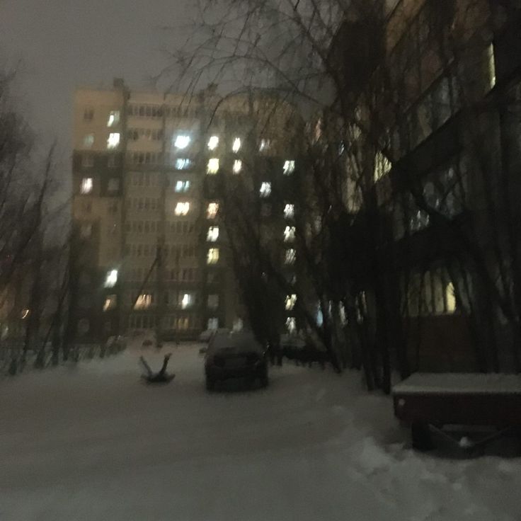 a snowy street with cars parked in front of tall buildings at night, lit by windows