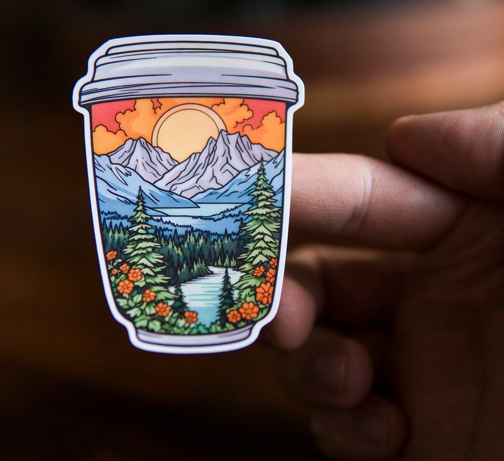 a hand holding up a sticker with mountains and trees in the background on it