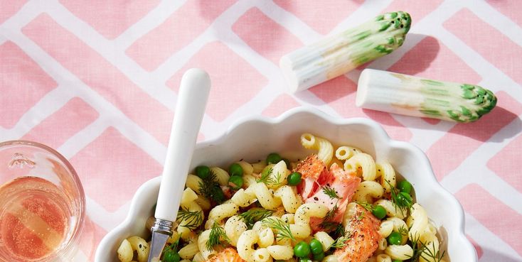 a bowl of pasta with shrimp and asparagus next to two wine glasses on a pink tablecloth