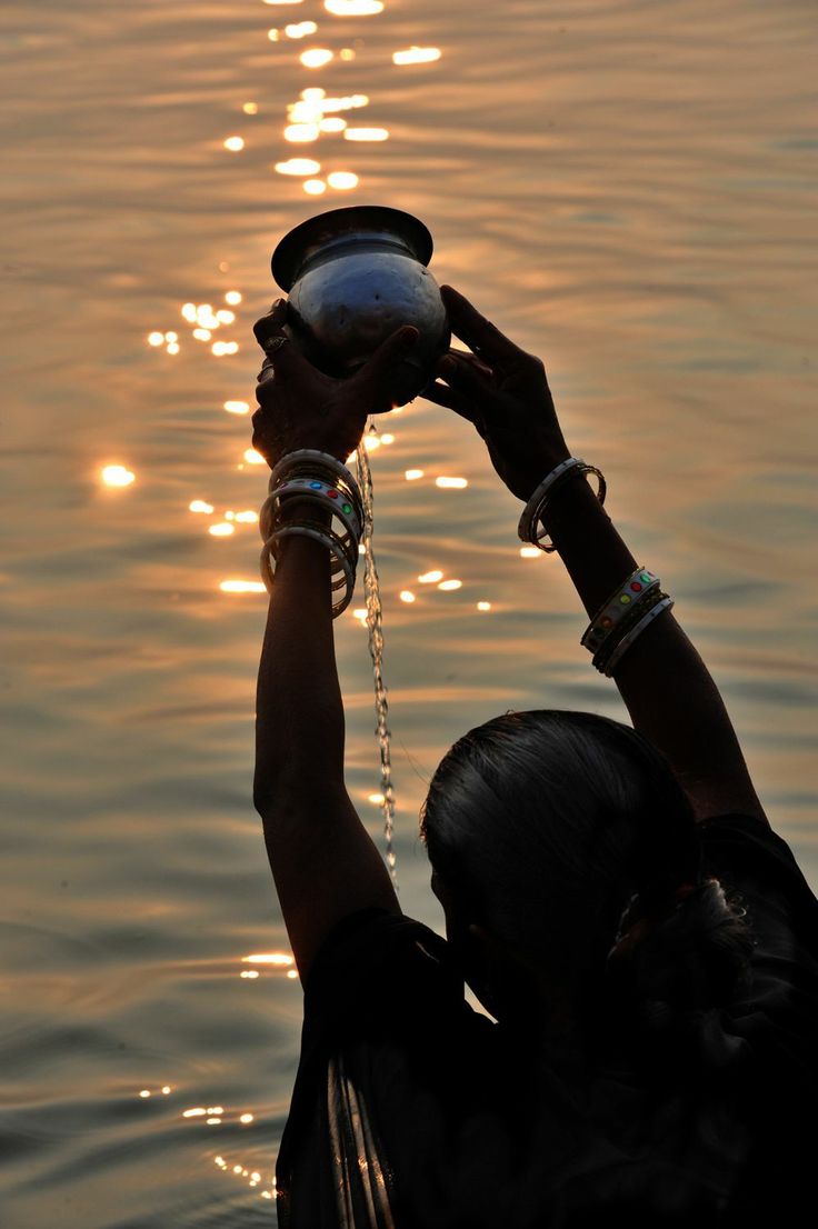 a woman holding up a bowl over her head while standing in the water at sunset