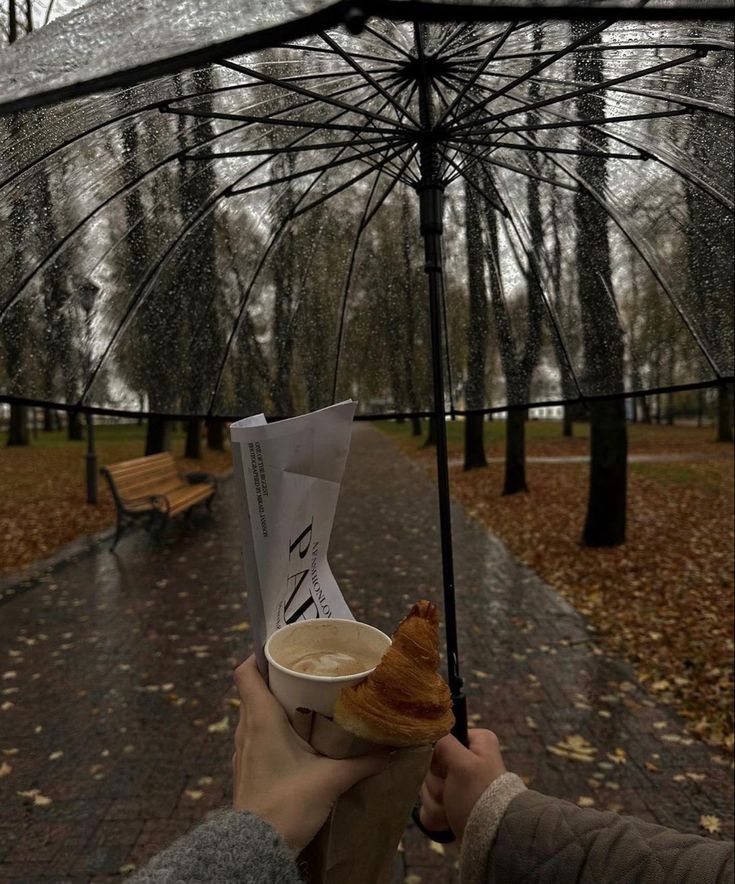 a person holding an umbrella over a cup of coffee and croissant in the rain