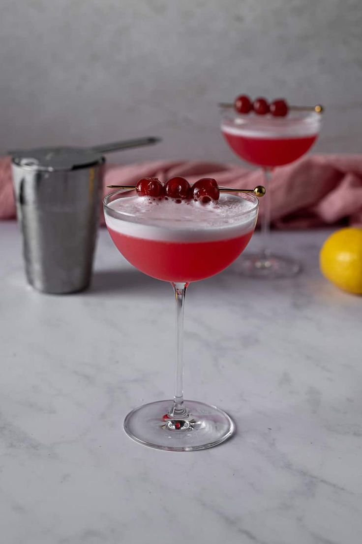 two martinis with cranberries sit on a marble table next to a cocktail shaker