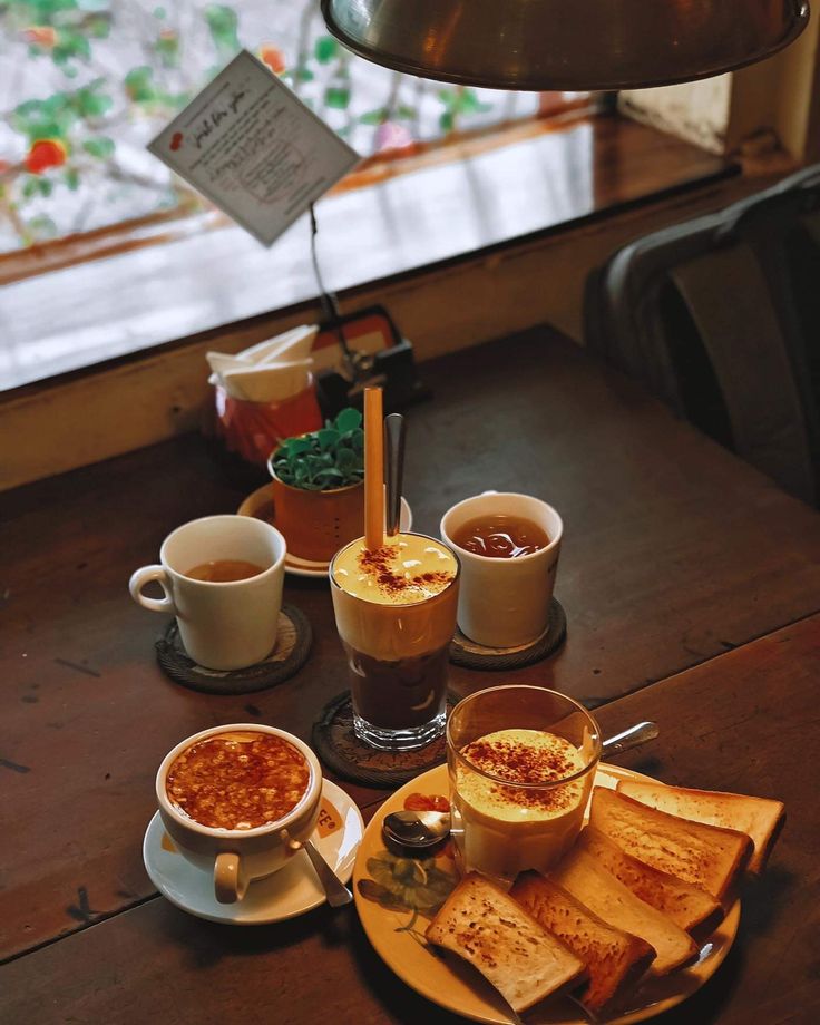 three cups of coffee sit on a wooden table with toast and other food in front of it