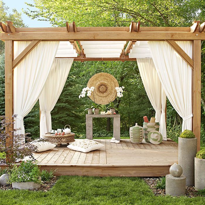 a wooden gazebo with white curtains and flowers on the ground in front of it