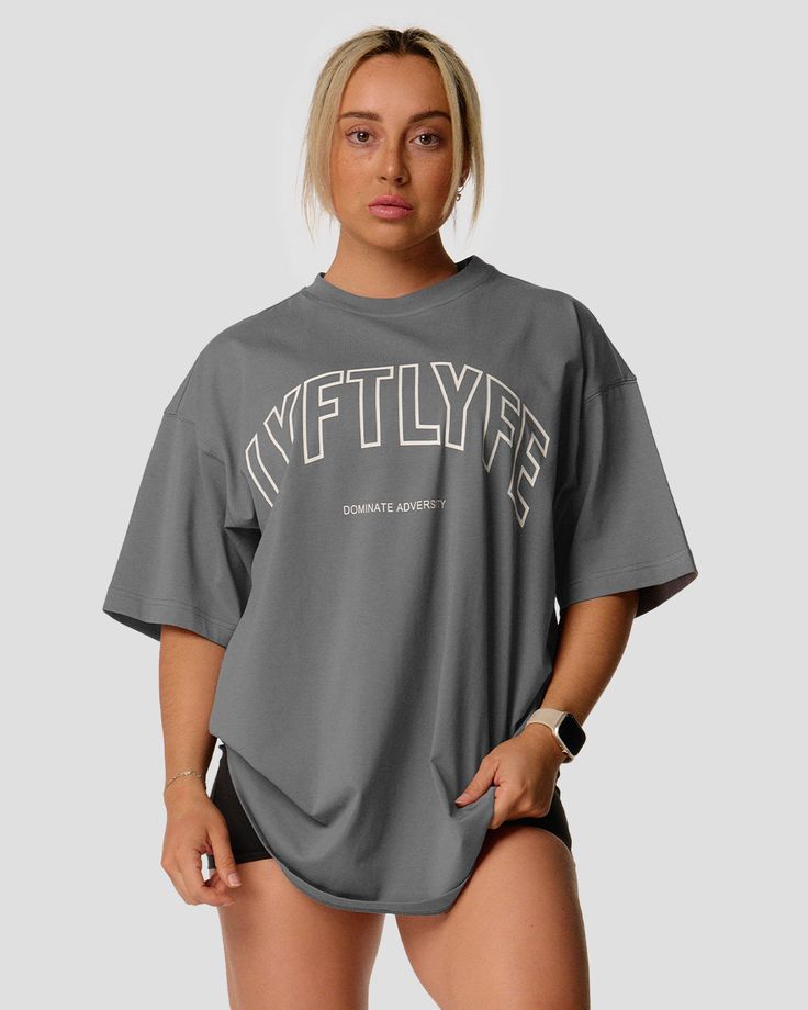 A female athlete is in a gray oversized T-shirt with a large lyftlyfe logo on the chest. Athleisure Tops With Letter Print And Drop Shoulder, Athleisure Letter Print Top With Drop Shoulder, Oversized Crew Neck T-shirt For Gym, Oversized Letter Print T-shirt For Gym, Oversized Graphic Tee For Sports, Oversized Letter Print Activewear, Sporty Boxy Fit T-shirt For Workout, Oversized Letter Print Athleisure Activewear, Oversized Moisture-wicking T-shirt For Athleisure