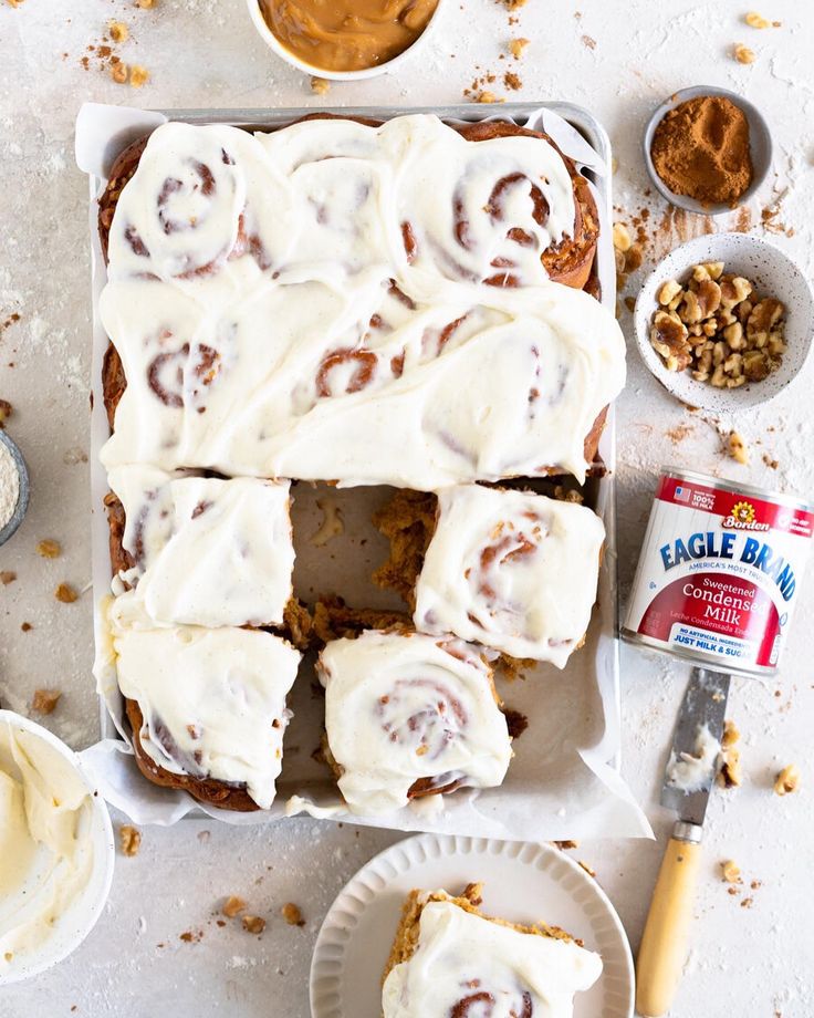 a pan filled with frosted cinnamon rolls next to bowls of nuts and sauces