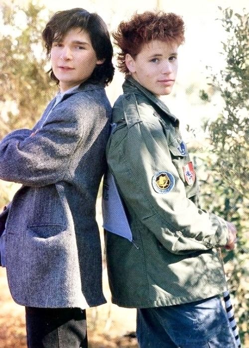 two young boys standing next to each other in front of trees and bushes, one with his arm around the other's shoulder