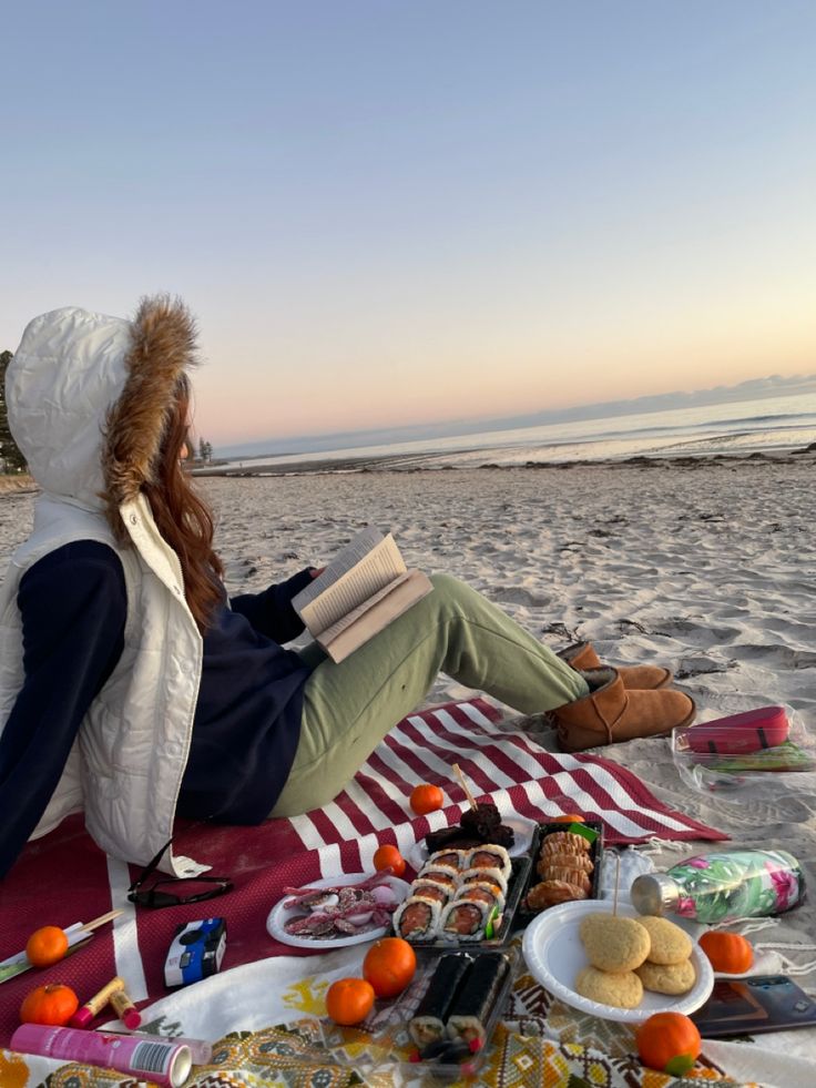 a woman sitting on top of a blanket next to an open book at the beach