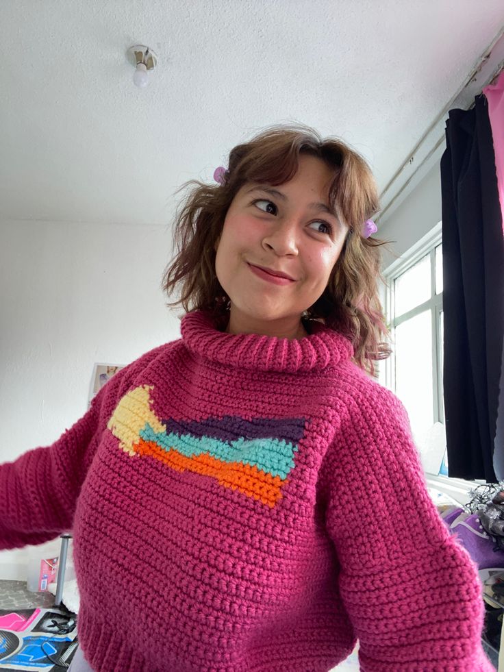 a woman wearing a pink knitted sweater standing in front of a window and smiling at the camera