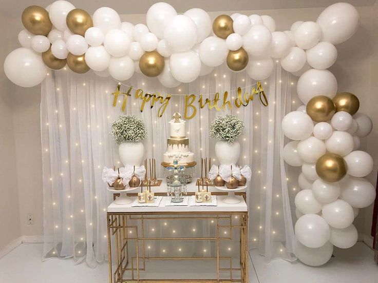 a table topped with white and gold balloons next to a cake on top of a table