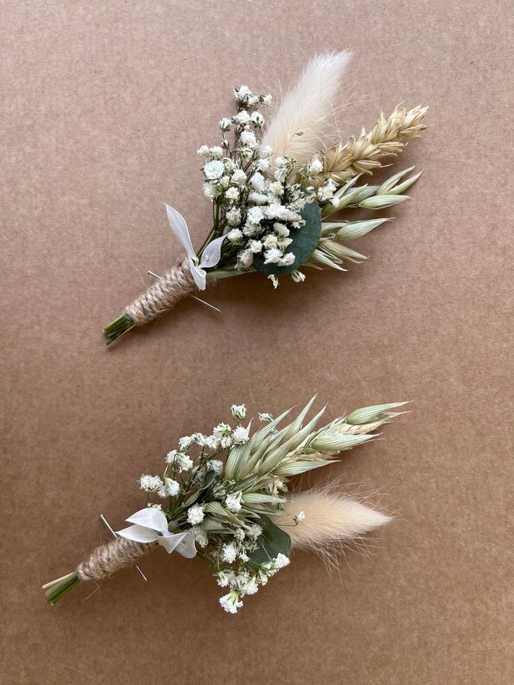 two boutonnieres with white flowers and feathers on brown paper, side by side