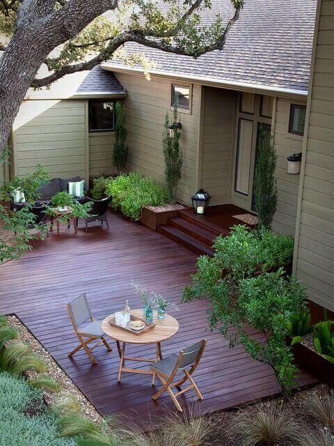 a wooden deck in front of a house surrounded by trees and bushes with chairs around it