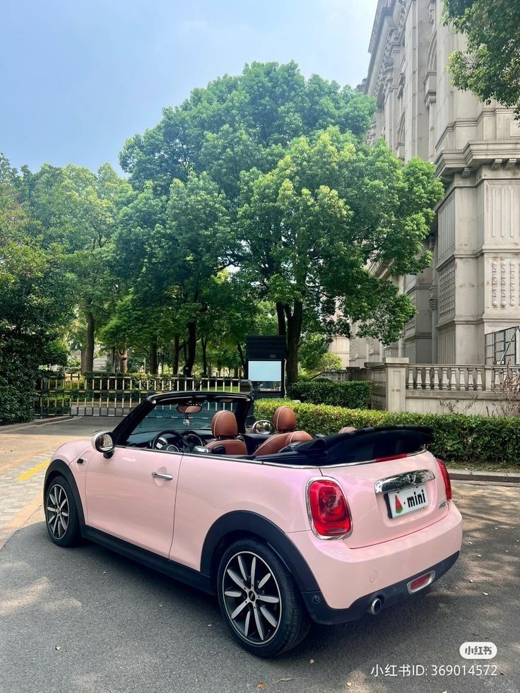 two people in a pink convertible car driving down the street with trees and buildings behind them