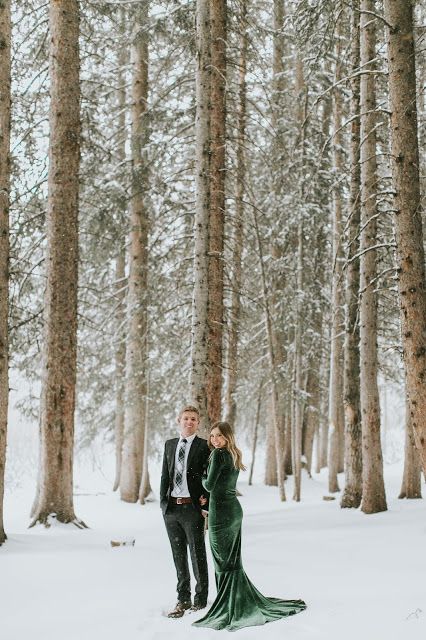 an engaged couple standing in the snow surrounded by trees