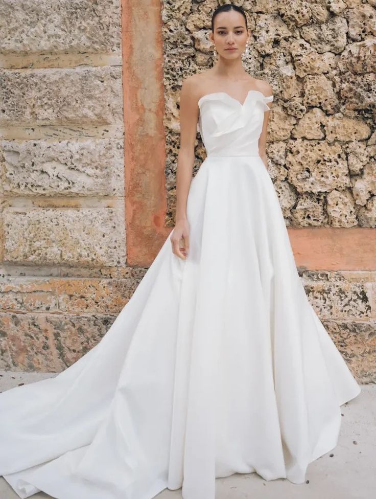 a woman standing in front of a stone wall wearing a white wedding dress with an asymmetrical neckline