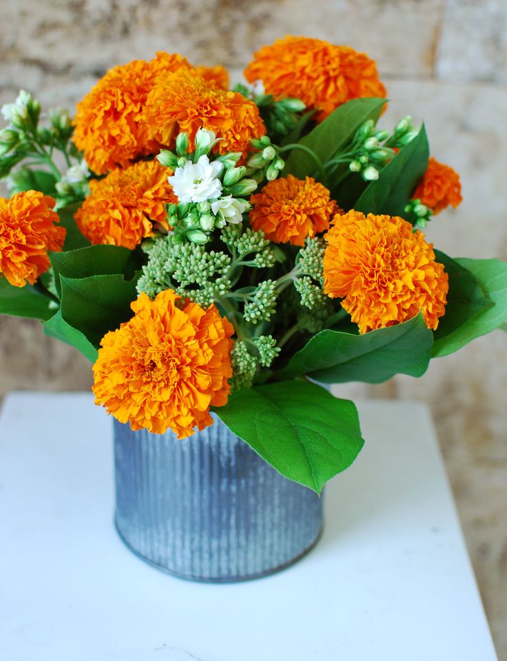 an orange and white flower arrangement in a blue tin vase on a table with green leaves