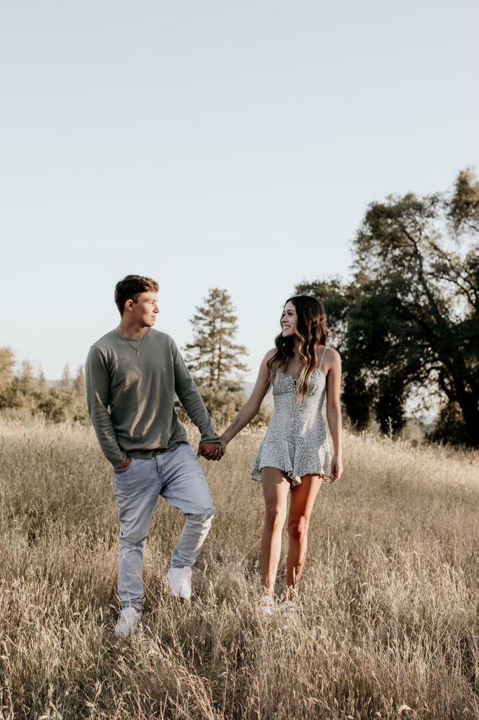 a man and woman holding hands walking through tall grass in an open field with trees behind them