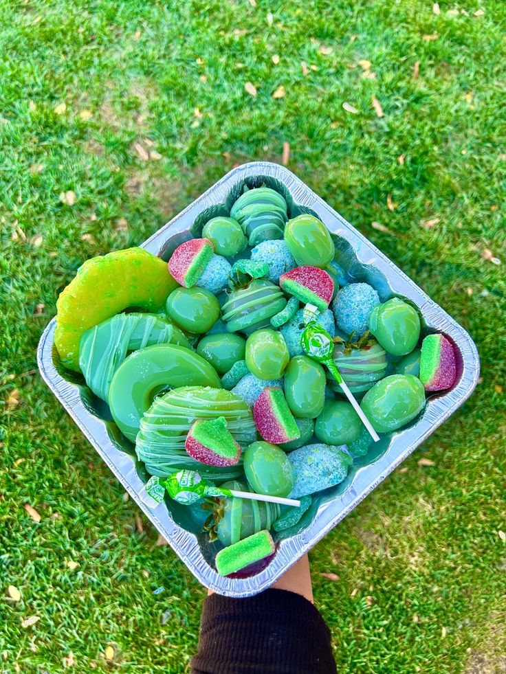 a person holding a container filled with green and purple candy on top of grass covered ground