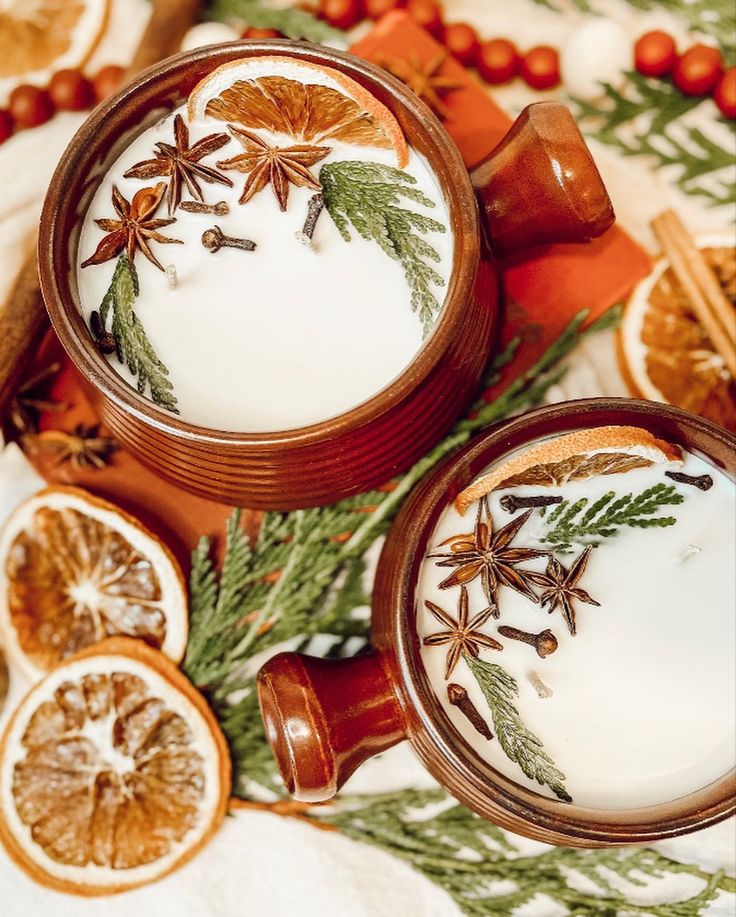 two mugs filled with white liquid surrounded by orange slices and cinnamon sticks on a table