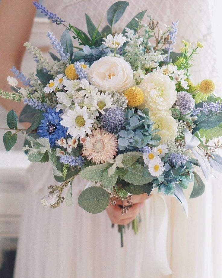 a woman holding a bouquet of flowers in her hands with greenery and blue thistles