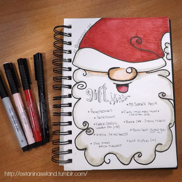 a spiral notebook with santa claus's face on it and three pens next to it