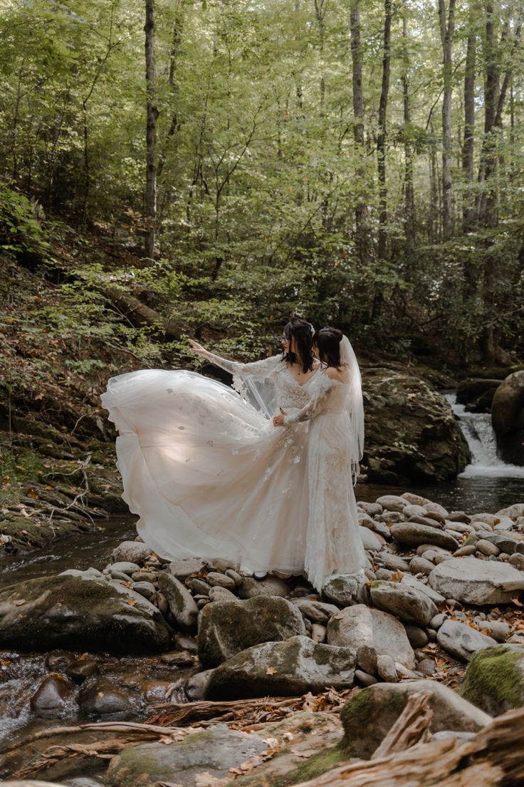 two brides are standing on rocks in the woods near a stream wearing long white dresses