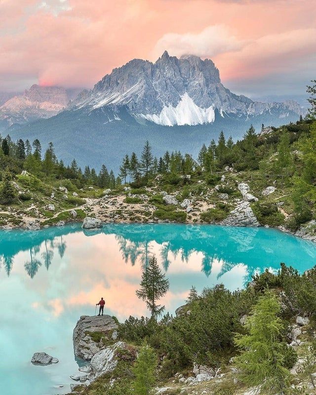 a person standing on top of a rock next to a lake with mountains in the background