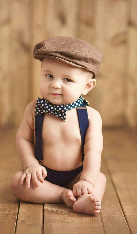 a baby sitting on the floor wearing a brown hat and blue overalls with a polka dot bow tie