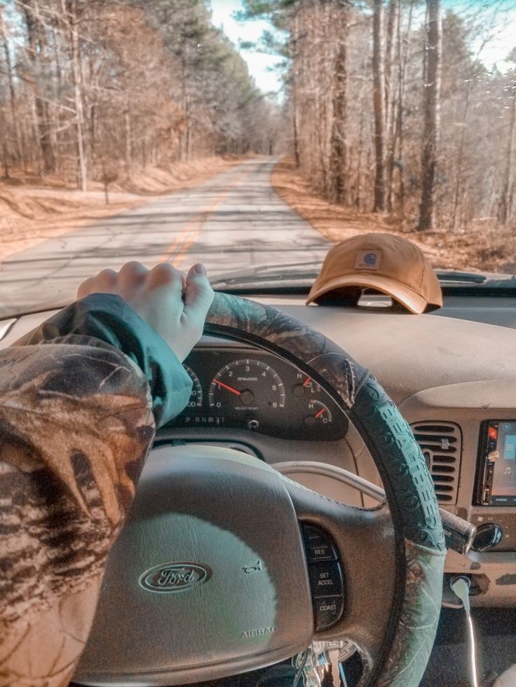 a person driving a car on a road with trees in the background and one hand on the steering wheel