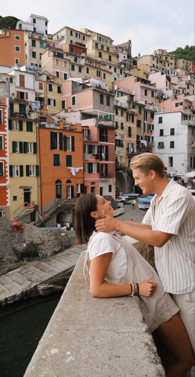 a man and woman standing next to each other on a bridge over water with buildings in the background