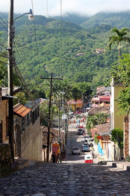 a cobblestone street with cars parked on both sides and mountains in the background