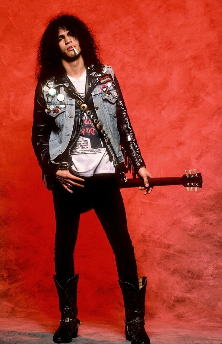 a man with long hair and leather jacket holding a guitar in front of a red wall