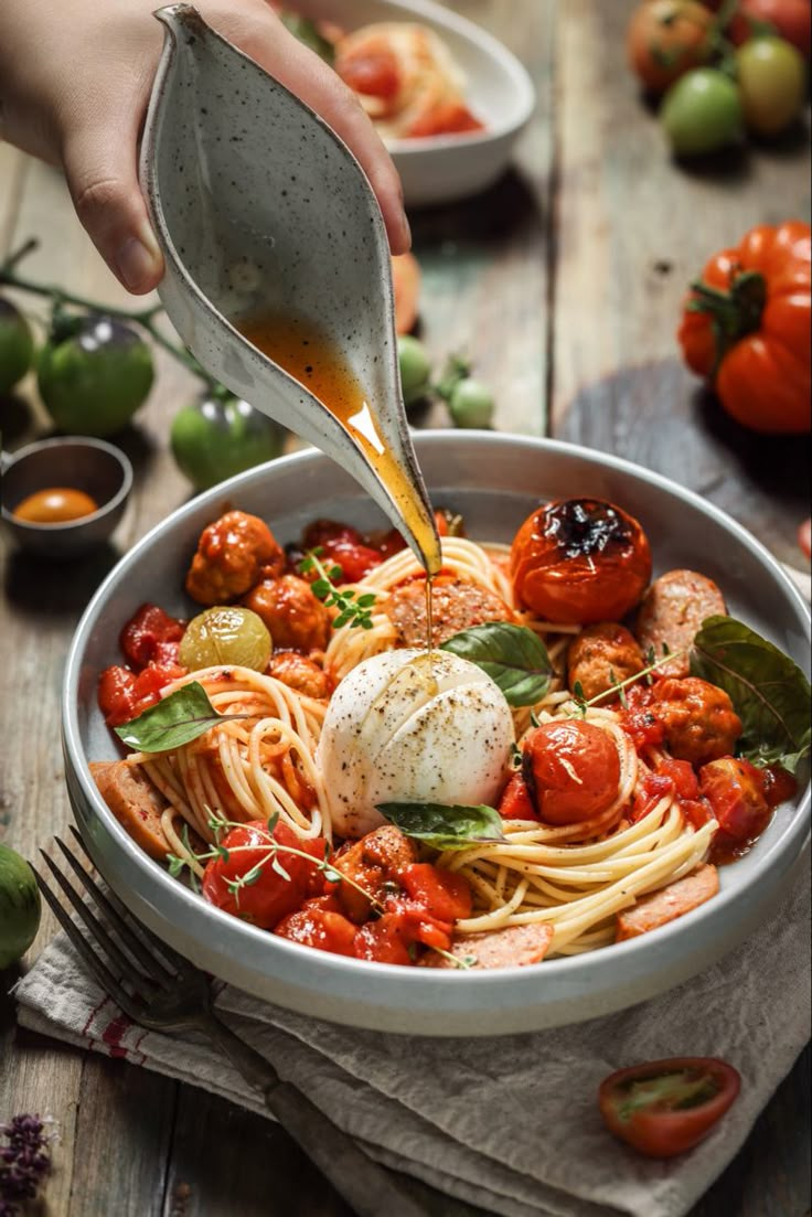 someone is pouring sauce over a bowl of spaghetti with tomatoes and basil on the side