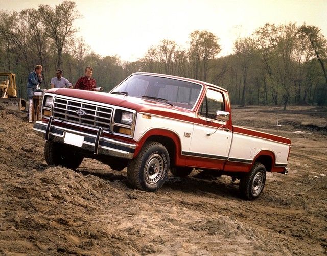 two men standing next to a red and white truck on a dirt road with trees in the background