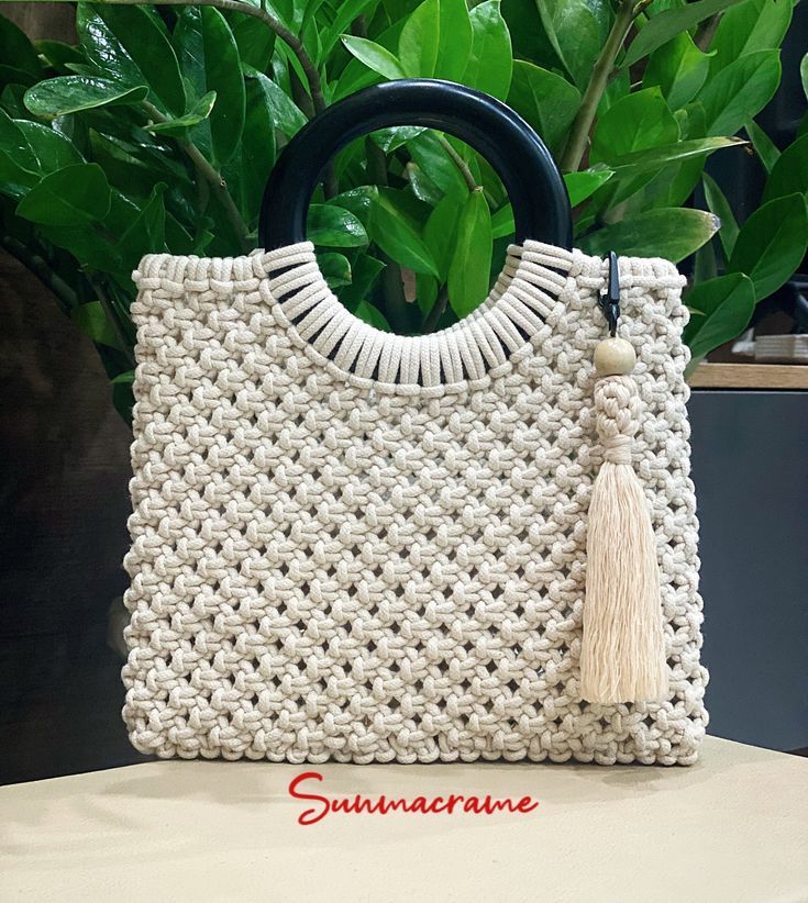 a white crocheted bag with tassels and a black handle sitting on top of a table