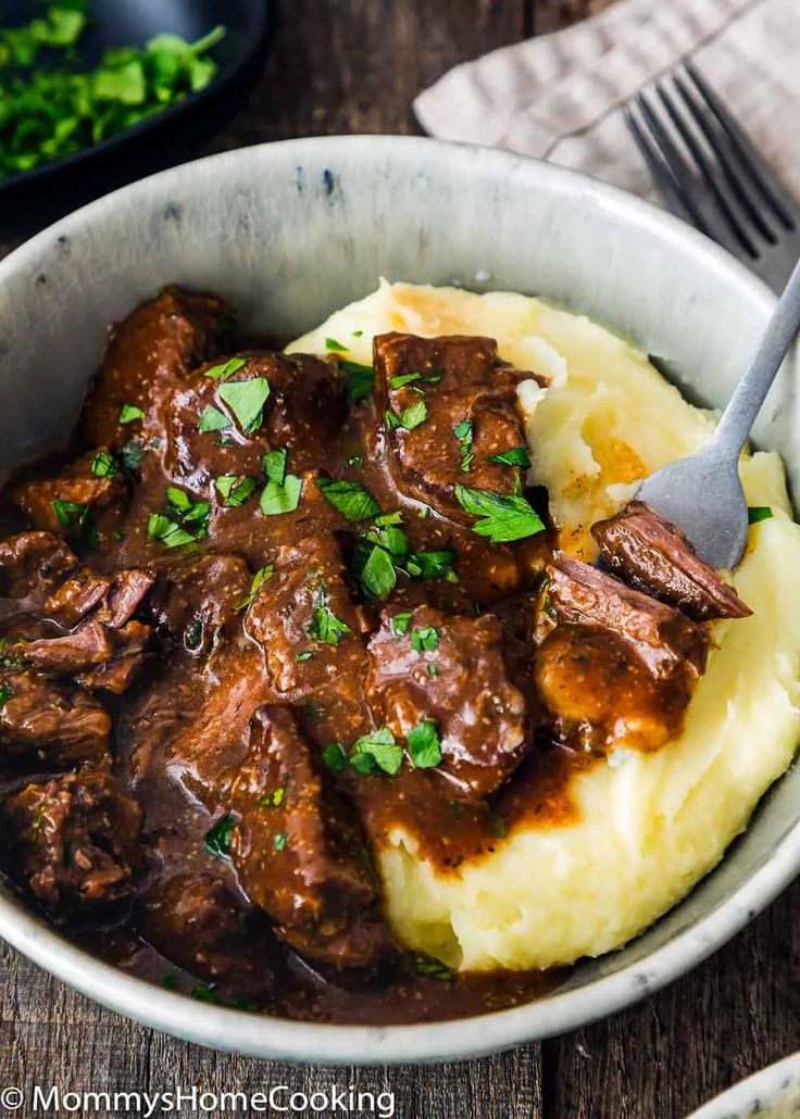 a bowl filled with meat and mashed potatoes on top of a wooden table next to a fork