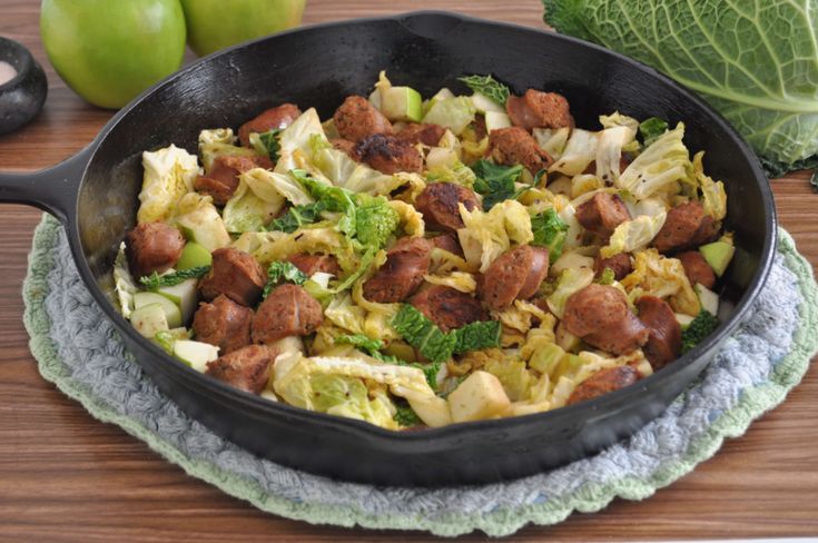 a skillet filled with meat and vegetables on top of a wooden table next to cabbage