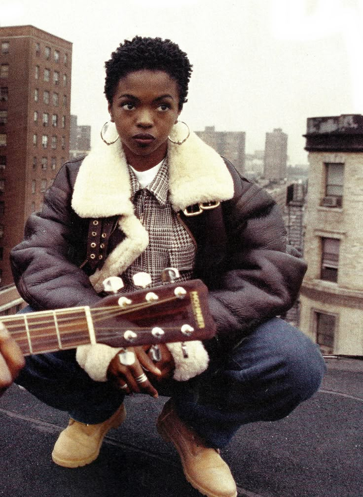 a man sitting on top of a roof with an acoustic guitar in his hand and buildings in the background