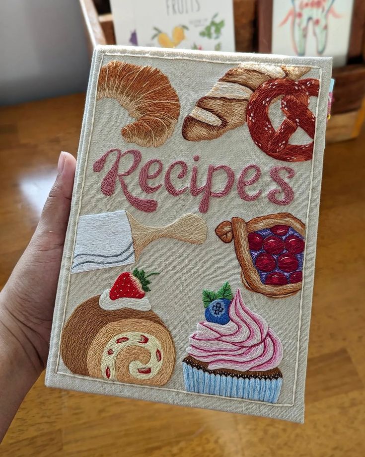 a hand holding up an embroidered book with pictures of food and pastries on it