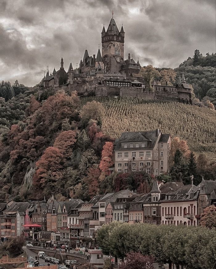 an old castle on top of a hill with autumn trees in the foreground and buildings below