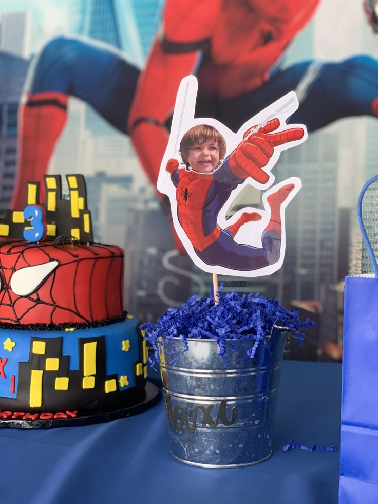 a spiderman birthday cake and cupcake topper on a table with blue streamers