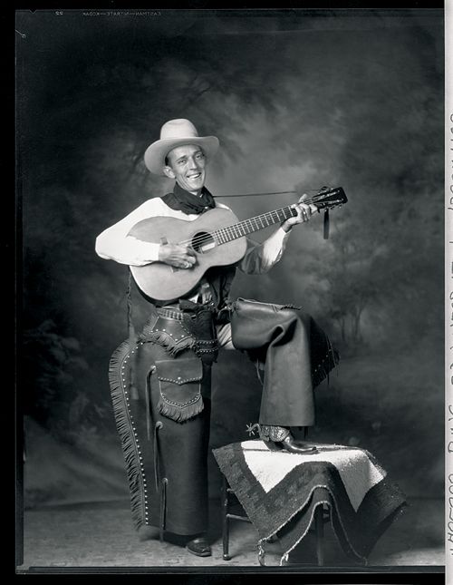 a black and white photo of a man with a guitar in his hand wearing a cowboy outfit