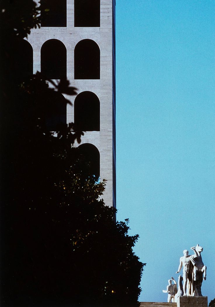 there is a clock tower in the distance with statues on it's sides and trees below