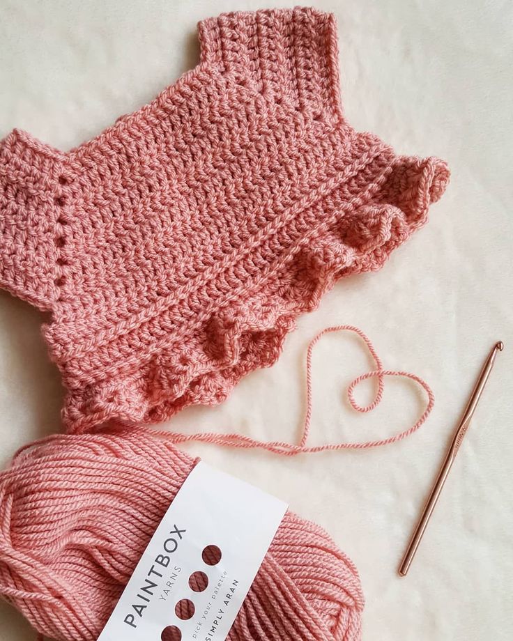 a pink knitted sweater next to a ball of yarn and knitting needles on a white surface