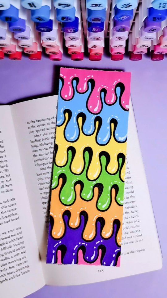 an open book next to some markers and pens on a purple table with books in the background