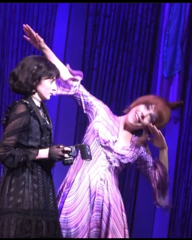 two women in dresses on stage with one holding her hand out and the other pointing