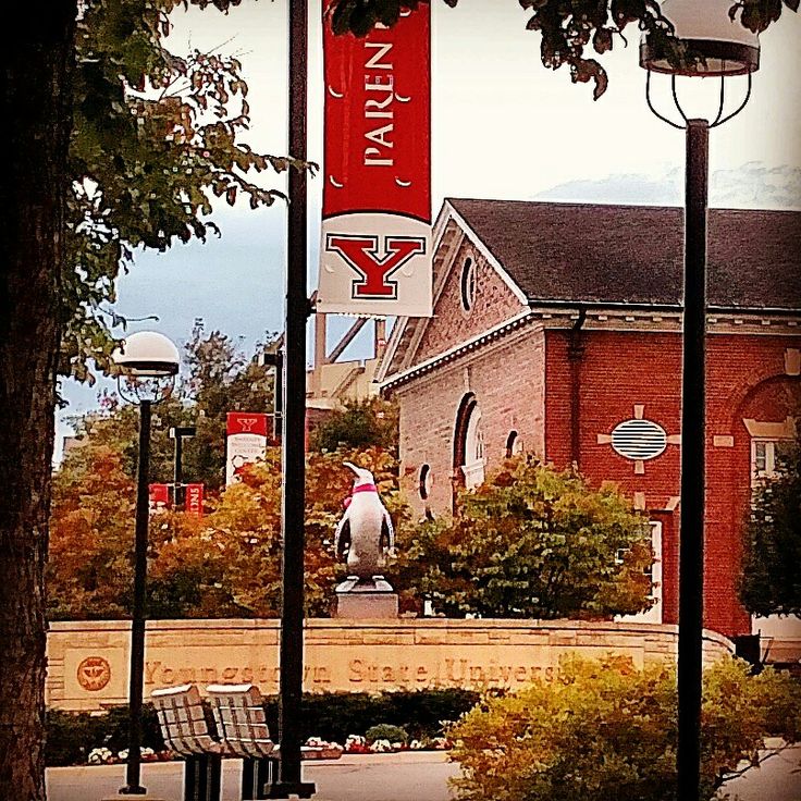 an old brick building with a large red sign hanging from it's side next to benches and trees