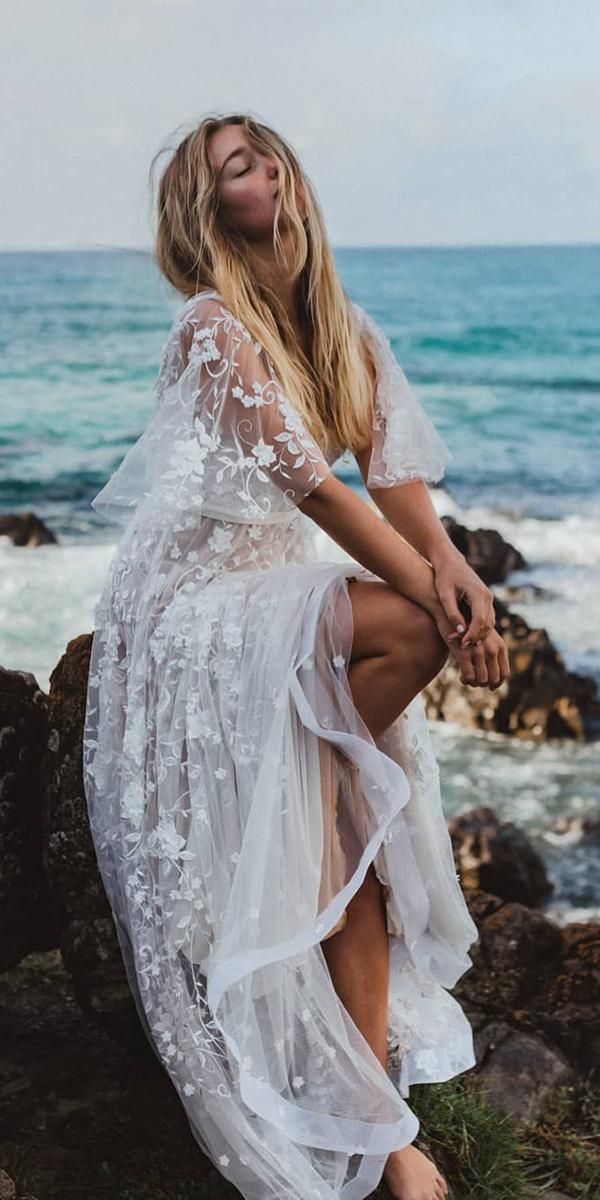 a woman is sitting on rocks near the ocean wearing a white dress with sheer sleeves